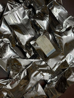 A pile of shiny silver resealable pouches, with one cream-colored label card visible in the center, photographed close up against a dark background.