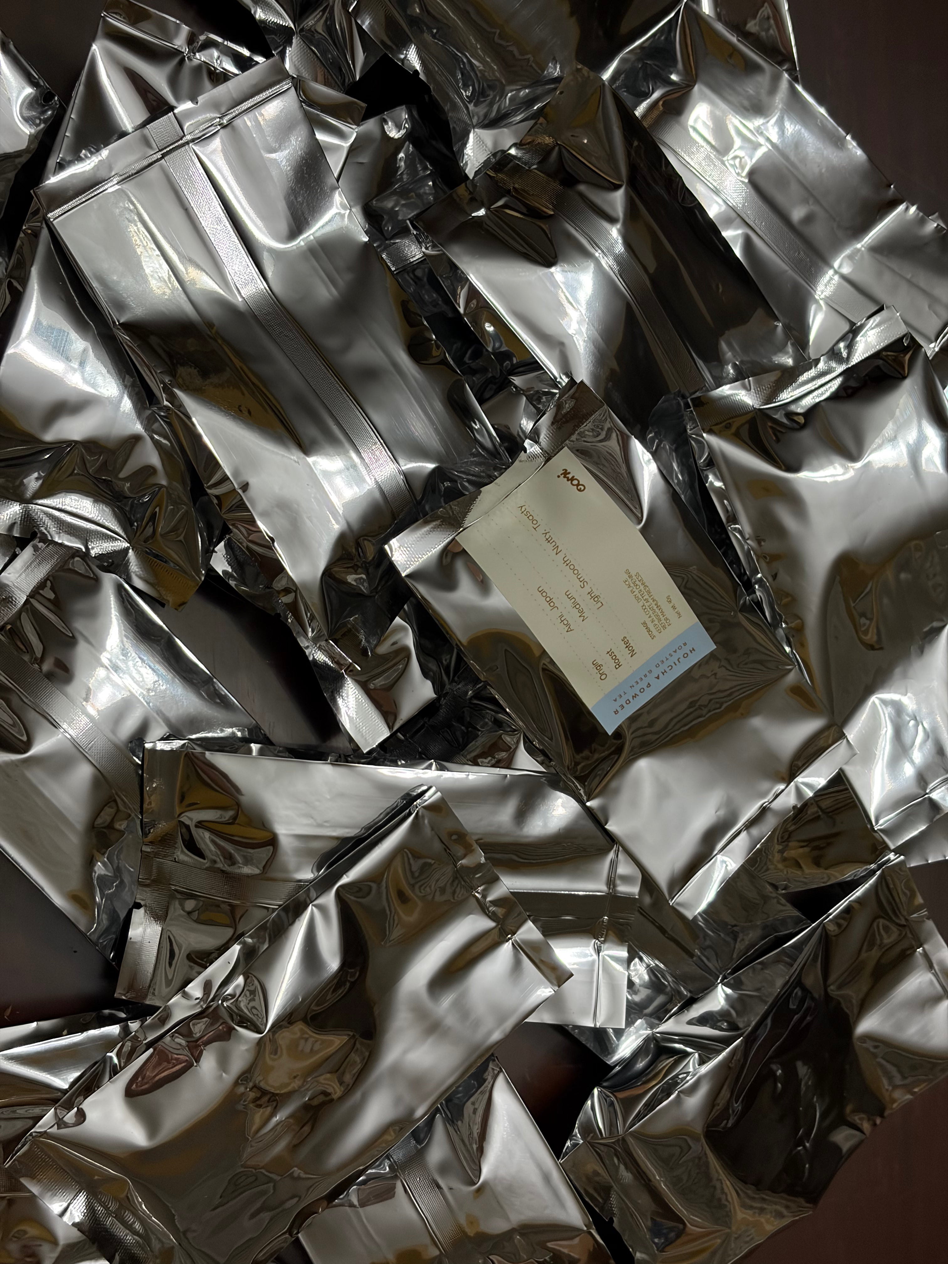 A pile of shiny silver resealable pouches, with one cream-colored label card visible in the center, photographed close up against a dark background.