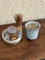 A ceramic tea set displayed on a wooden surface, including a small cup with a brown base, a matching bowl, and a bamboo whisk.