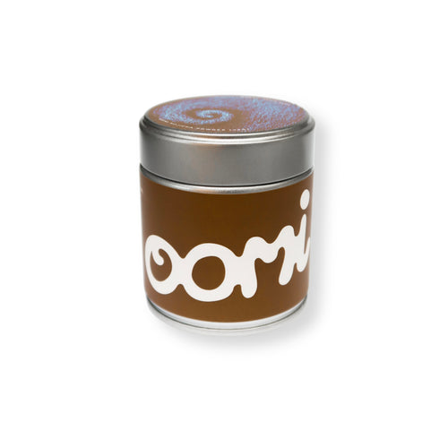 A cylindrical metal tin candle with a brown paper label featuring the white stylized text 'oomi' and a silver lid, shown on a plain white background. This metal tin contains 40 grams of Hojicha Powder.